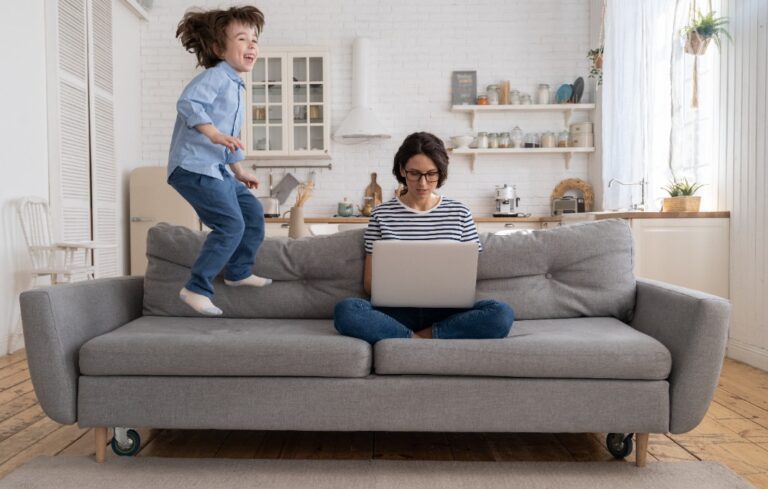 Focused young mother freelancer wear glasses sitting on couch working on laptop at home, trying to concentrate and not be distracted, hyperactive little son jumping on sofa and drawing