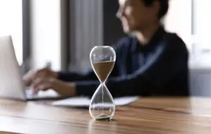 Close up hourglass measuring time, standing on wooden office table, businesswoman working on background,