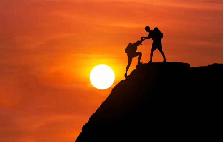 silhouette mountaineer gives helping hand his friend to climb high cliff mountain together.