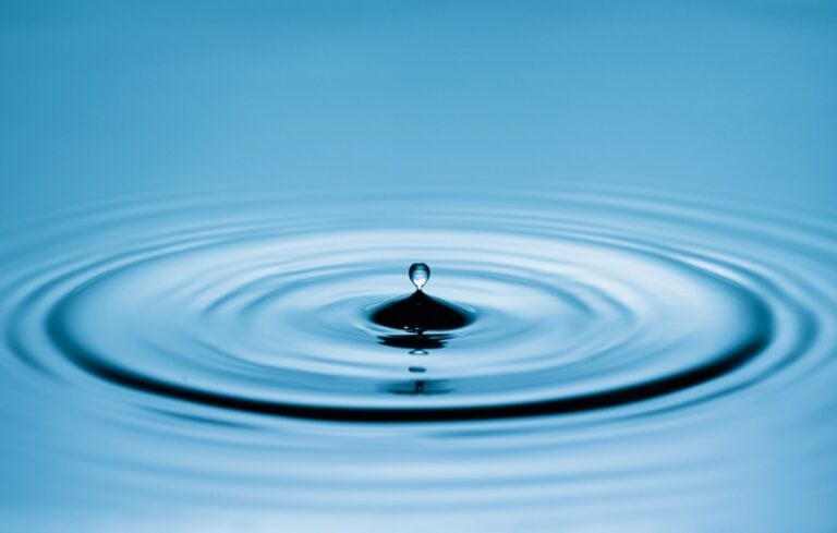Close up of a blue water drop macro creating concentric ripples on a calm water surface.