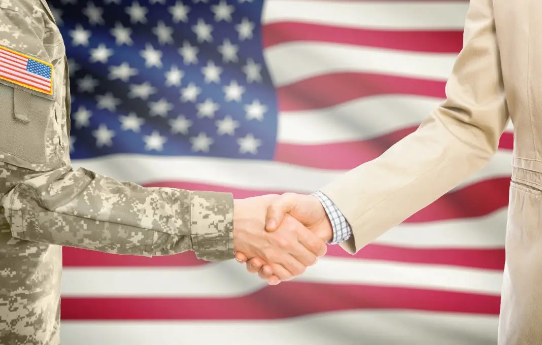 USA military man in uniform and civil man in suit shaking hands with adequate national flag on background - United States of America